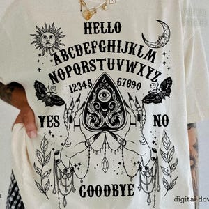 May include: Cream t-shirt featuring a black Ouija board design. The graphic includes the alphabet, numbers, and the words "Hello," "Yes," "No," and "Goodbye." Decorative elements include a sun, moon, butterflies, and hands.