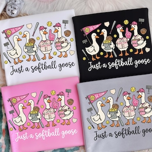 Op de afbeelding: Vier t-shirts met een softbalthema. Elk shirt heeft een cartoon gans met een honkbalpet en softbal-gerelateerde items. De tekst "Just a softball goose" staat eronder. De shirts zijn in wit, zwart, roze en grijs.