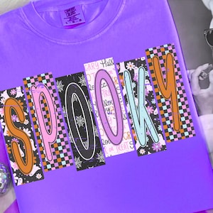 Może przedstawiać: Fioletowa koszulka Comfort Colors z napisem "SPOOKY" w kolorowych, wzorzystych literach. Litery są pomarańczowe, różowe, czarne, jasnoniebieskie i pomarańczowe, z motywami Halloween. W tle magazyn z kobietą w okularach przeciwsłonecznych.