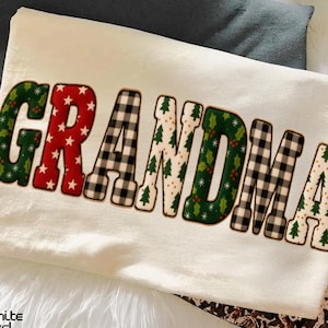 Könnte beinhalten: Cremefarbenes T-Shirt mit dem Wort "GRANDMA" in einem festlichen Design. Die Buchstaben zeigen Muster aus roten Sternen, schwarz-weißem Büffelkaromuster, grünem Stechpalmen und Weihnachtsbäumen. Im Hintergrund ist ein dunkelgraues T-Shirt zu sehen.