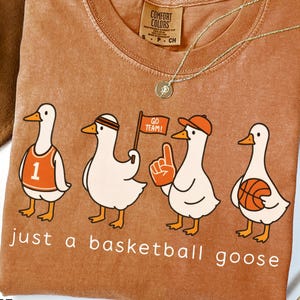 May include: A rust-coloured Comfort Colors t-shirt with a cartoon design of four geese dressed for basketball. The geese wear a jersey, headband, hat, and hold a flag and basketball. The text "just a basketball goose" is below the geese.