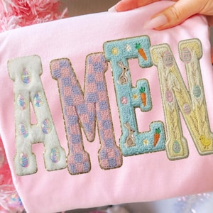 Könnte beinhalten: Ein hellrosa Shirt mit dem Wort "AMEN" in bunten, dekorativen Buchstaben gestickt. Die Buchstaben zeigen Ostermotive, darunter Eier, Hasen und Karotten. Das Shirt wird vor einem rosa Weihnachtsbaum gehalten.
