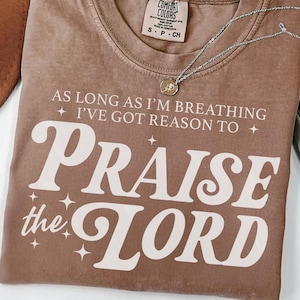 Può includere: Una maglietta marrone Comfort Colors con la scritta bianca "AS LONG AS I'M BREATHING I'VE GOT REASON TO PRAISE the LORD". Una collana d'argento con un ciondolo è sulla maglietta. La maglietta è piegata.