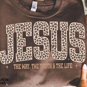May include: A brown sweatshirt featuring the word "JESUS" in large leopard print lettering. Underneath, the phrase "THE WAY. THE TRUTH & THE LIFE" is printed in a smaller, brown font. The sweatshirt is held up by two hands.