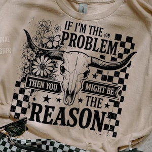 May include: Beige t-shirt with a graphic featuring a bull skull, flowers, and the text "IF I'M THE PROBLEM THEN YOU MIGHT BE THE REASON." A black and white checkered fanny pack, sunglasses, and a scrunchie are also visible.