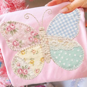 Può includere: Un cuscino rosa chiaro con un'applicazione a farfalla. Le ali della farfalla sono realizzate con tessuti floreali, a quadretti e a pois, con dettagli in pizzo. Il corpo della farfalla è cucito con un filo decorativo.