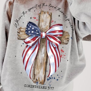 K&ouml;nnte beinhalten: Grauer Pullover mit einem Holzkreuz und einer rot-wei&szlig;-blauen Schleife. Das Kreuz ist mit Sternen und dem Text "Where the spirit of the Lord there is freedom" und "Corinthians 3:17" verziert.