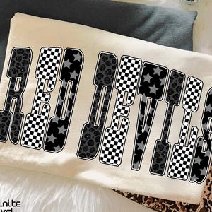 May include: Cream-colored t-shirt with the words "RED DEVILS" in a bold, stylized font. The letters feature a mix of patterns, including leopard print, checkered, and star designs. The shirt is folded on a gray surface.