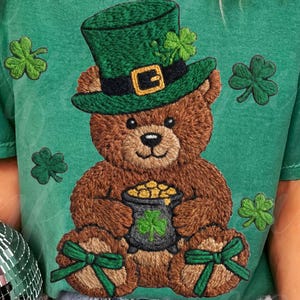 May include: Green t-shirt featuring a brown teddy bear wearing a green top hat with a gold buckle and shamrock accents. The bear holds a pot of gold and is surrounded by shamrock designs. The shirt has a vintage, worn look.