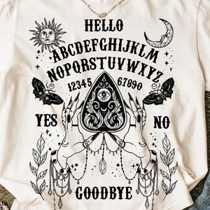May include: Cream-coloured t-shirt with a black Ouija board design. The graphic includes the alphabet, numbers, sun, moon, butterflies, and the words "Hello", "Yes", "No", and "Goodbye". The design is centered on the front of the shirt.