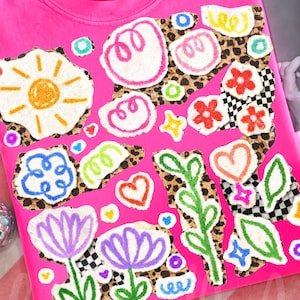 Op de afbeelding: Een felroze Comfort Colors t-shirt met een kleurrijk, grillig ontwerp. Het ontwerp bevat een zon, bloemen, harten en andere vormen met een luipaardprint en dambordpatroon. De tekst "Better Late The" is zichtbaar op de achtergrond.