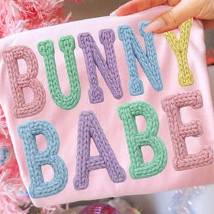 Może przedstawiać: Jasnoróżowa koszulka z napisem "BUNNY BABE" w kolorowych, teksturowanych literach. Litery są w pastelowych odcieniach różu, zieleni, błękitu, fioletu i żółci. Koszulka jest trzymana przez dłoń, z różową choinką w tle.