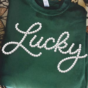 以下が含まれることがあります： 白いイミテーションパールで「Lucky」の文字が書かれた濃い緑色のスウェットシャツ。スウェットシャツは折りたたまれ、人が持っています。背景には模様のある生地とキャンドルが含まれています。