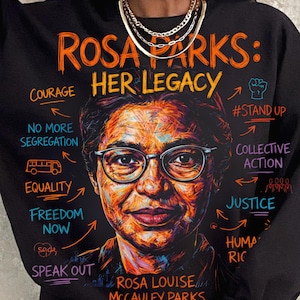 May include: Black sweatshirt with a vibrant portrait of Rosa Parks. The text "ROSA PARKS: HER LEGACY" is displayed above the portrait. Other text includes "COURAGE", "EQUALITY", "FREEDOM NOW", and "SPEAK OUT".
