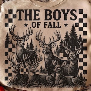 Könnte beinhalten: Beiges Sweatshirt mit einer Grafik, die sechs Hirsche mit großen Geweihen vor dem Hintergrund von Bäumen und Schachbrettmustern zeigt. Der Text "THE BOYS OF FALL" ist oben in fetter, schwarzer Schrift gedruckt.