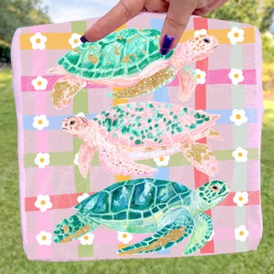 May include: A decorative rectangular item featuring three sea turtles in shades of green, pink, and blue, each with gold accents. The background is a pink plaid pattern with white flower accents. The item is held by a person.