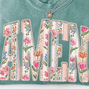 Puede incluir: Una camiseta Comfort Colors verde mar con la palabra "AMEN" en letras grandes con estampado floral. Las letras están delineadas con pequeñas cuentas perladas. Un collar plateado con un pequeño colgante descansa sobre la camisa.