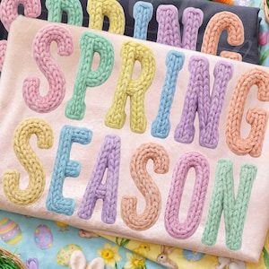May include: Two t-shirts with the words "SPRING SEASON" and "SPRING" in pastel knitted letters. The shirts are on a colorful Easter-themed background with eggs, carrots, and a bunny.