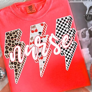 May include: Bright coral-colored t-shirt with the word "Nurse" in a white script font. The word is superimposed on three lightning bolt designs with leopard print, cherry, and checkered patterns. The shirt has a crew neck.