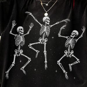 Può includere: T-shirt nero con grafica di tre scheletri bianchi danzanti su sfondo stellato. Gli scheletri sono raffigurati in una posa da balletto. La maglietta ha un aspetto vintage e usurato.