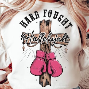 Może przedstawiać: Biała bluza z nadrukiem graficznym przedstawiającym drewniany krzyż, różowe rękawice bokserskie oraz napisy "HARD FOUGHT Hallelujah". Projekt zawiera małe krzyże i linie rozbłysków.