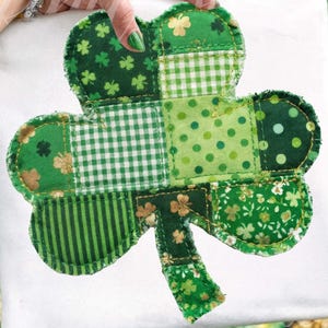 Könnte beinhalten: Ein Patchwork-Kleeblatt in Grüntönen mit verschiedenen Mustern wie Punkten, Streifen und floralen Drucken. Das Kleeblatt wird vor einem weißen Hintergrund präsentiert, der die detaillierten Nähte und Stofftexturen hervorhebt. Das Design ist wahrscheinlich für St. Patrick's Day.