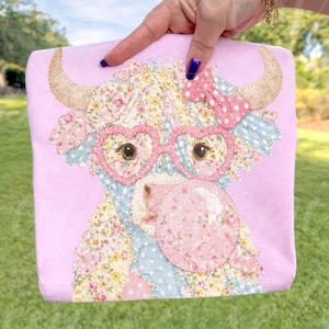May include: A pink tote bag featuring a patchwork cow design. The cow has heart-shaped glasses, a pink bow, and floral and patterned patches. The horns are tan, and the background is a grassy area.