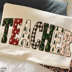 Könnte beinhalten: Cremefarbenes Langarmshirt mit dem Wort "TEACHER" in einem festlichen, gemusterten Design. Die Buchstaben zeigen Stechpalmen, Sterne, Weihnachtsbäume und ein schwarz-weißes Büffelkaromuster. Im Hintergrund ist ein dunkelgraues Shirt zu sehen.