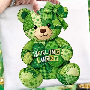 May include: A white t-shirt featuring a green patchwork teddy bear holding a shamrock with the words "FEELING LUCKY". The bear has a green bow and is made of various green patterned fabrics.