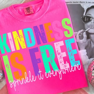 以下が含まれることがあります： 鮮やかなピンク色のTシャツ。カラフルでかすれた文字で「KINDNESS IS FREE」と書かれています。その下に「sprinkle it everywhere」というフレーズが筆記体で書かれています。クルーネックで半袖です。
