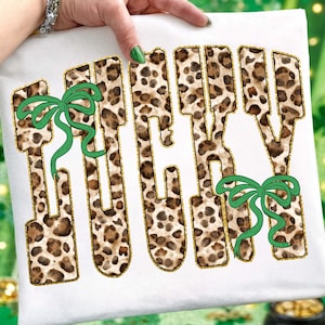 May include: White t-shirt with the word "LUCKY" in a leopard print design outlined in gold glitter. Two green bows are placed on the shirt. The shirt is held up by a person.