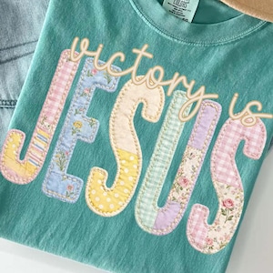 May include: A teal t-shirt with the words "victory is JESUS" in a patchwork design. The shirt is paired with light-wash jeans, a gold necklace, and a tan hat with a brown band. Dried flowers are also visible.
