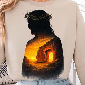 Könnte beinhalten: Beiges Sweatshirt mit einer Silhouette einer Figur mit einer Dornenkrone und einem inneren Bild eines Sonnenuntergangs über einem Grab. Die Grafik ist in Orange, Gelb und Schwarz gehalten. Das Sweatshirt hat eine neutrale Farbe.