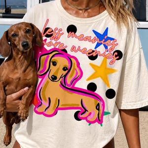 Puede incluir: Camiseta color crema con una ilustración de perro salchicha de dibujos animados y el texto "less meanies, more weenies". El diseño incluye una estrella amarilla, una estrella azul y lunares negros. Un perro salchicha real se sostiene frente a la camiseta.
