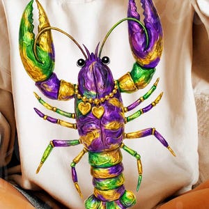 May include: Cream-colored sweatshirt featuring a vibrant, detailed illustration of a Mardi Gras lobster. The lobster is adorned with purple, green, and gold accents, and a heart-shaped necklace. The design is centered on the front of the shirt.