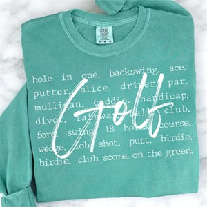May include: A teal green t-shirt with white text that reads "hole in one. backswing. ace. putter. slice. driver. par. mulligan. caddie. handicap. divot. fairway. 18 holes. course. wedge. lob shot. putt. birdie. birdie. club. score. on the green." The word "Golf" is written in large white letters over the text.