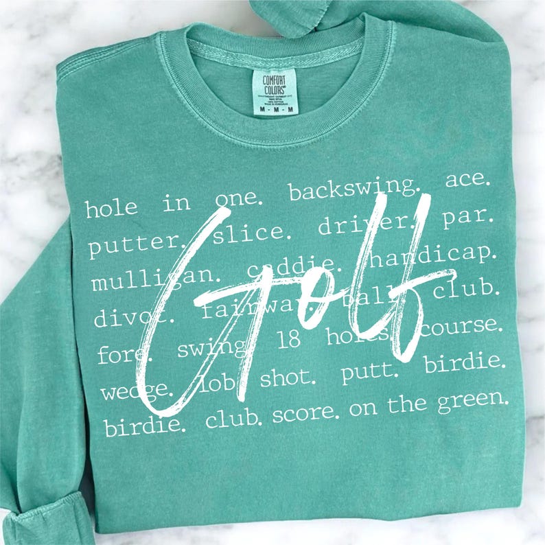 May include: A teal green t-shirt with white text that reads "hole in one. backswing. ace. putter. slice. driver. par. mulligan. caddie. handicap. divot. fairway. 18 holes. course. wedge. lob shot. putt. birdie. birdie. club. score. on the green." The word "Golf" is written in large white letters over the text.