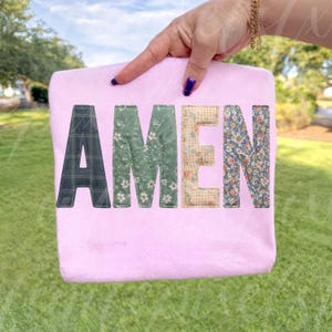 May include: A light pink tote bag with the word "AMEN" in patchwork letters. The letters are in various patterns, including plaid, floral, and gingham. The bag is held up by a hand with blue nail polish.