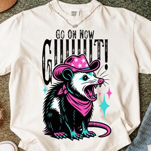 May include: Cream-colored t-shirt featuring a graphic of an opossum wearing a pink cowboy hat and bandana. The opossum has an open mouth with visible teeth. The text "GO ON NOW GIT IT!" is printed above the opossum.
