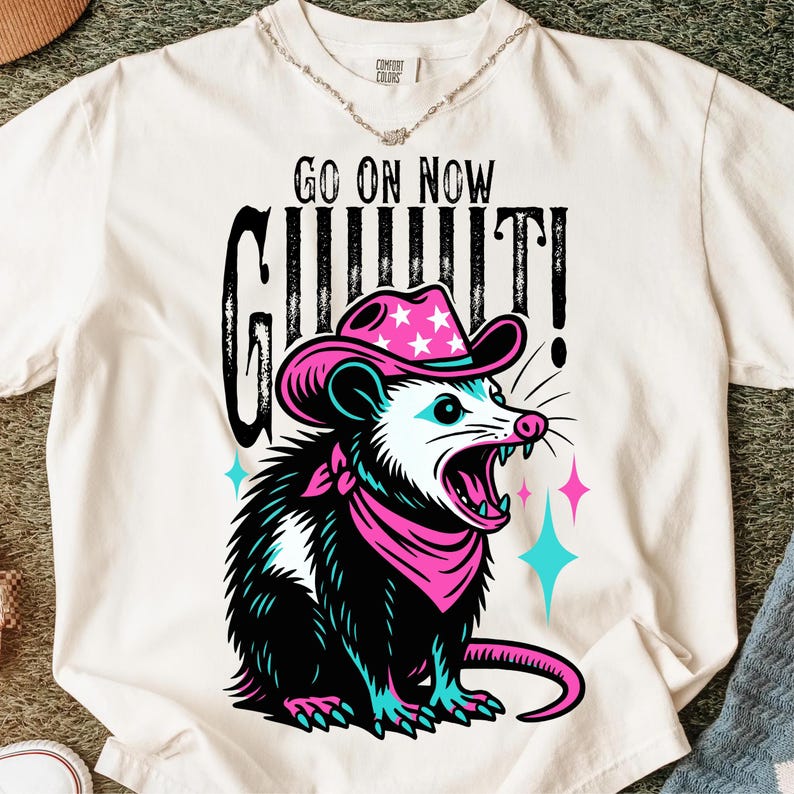 May include: Cream-colored t-shirt featuring a graphic of an opossum wearing a pink cowboy hat and bandana. The opossum has an open mouth with visible teeth. The text "GO ON NOW GIT IT!" is printed above the opossum.