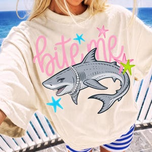 May include: Cream-colored t-shirt with the text "Bite Me" in pink script, featuring a cartoon shark wearing a pearl necklace. The shark is gray with white teeth and is surrounded by blue and green star accents.