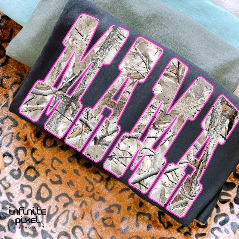 May include: A folded black sweatshirt with the word "MAMA" in large, bold letters. The letters have a camouflage pattern with a pink outline. The sweatshirt is resting on a leopard print surface.