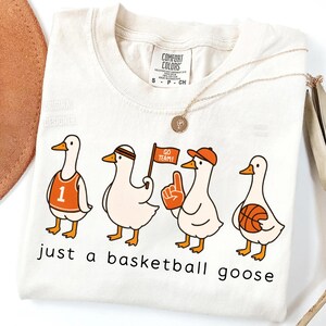 May include: Off-white t-shirt with a cartoon illustration of four geese in basketball fan attire. The geese are wearing a jersey, headband, hat, and holding a flag, foam finger, and basketball. The text "just a basketball goose" is printed below.