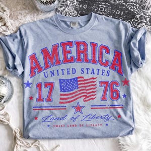 May include: Light blue t-shirt with a distressed red and blue graphic. The graphic includes the word "AMERICA" above "UNITED STATES", the number "1776", a US flag, and the phrases "Land of Liberty" and "Sweet Land of Liberty".