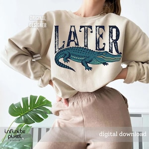 Pode incluir: Uma camisa de moletom bege com a palavra "LATER" impressa em preto e um gr&aacute;fico de um jacar&eacute; azul.