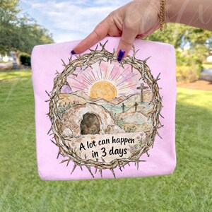 May include: A light pink t-shirt featuring a watercolor design of a sunrise over a desert landscape with a tomb and crosses, framed by a crown of thorns. The text "A lot can happen in 3 days" is below the image.