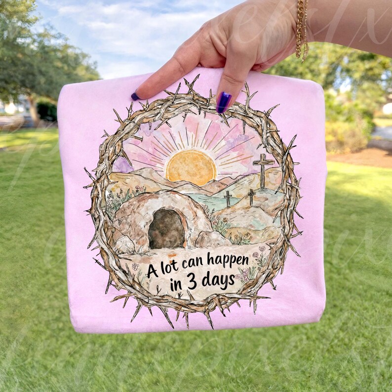 May include: A light pink t-shirt featuring a watercolor design of a sunrise over a desert landscape with a tomb and crosses, framed by a crown of thorns. The text "A lot can happen in 3 days" is below the image.
