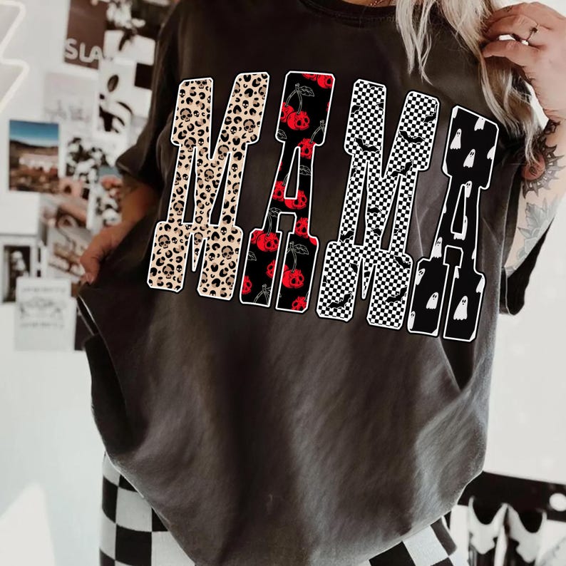 May include: Dark gray t-shirt with the word "MAMA" in a bold, decorative font. The letters feature various patterns: leopard print, red cherries, black and white checkerboard, and ghosts. The shirt is a casual, oversized style.