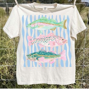 May include: Cream-colored t-shirt featuring three colorful fish illustrations. The fish are in shades of green, pink, and blue, set against a light blue wavy background. The shirt is hanging on a clothesline. The text "COMFORT COLORS" is printed on the upper right.