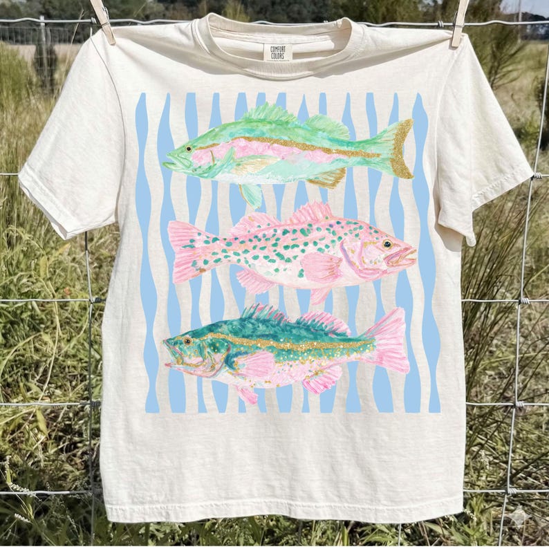 May include: Cream-colored t-shirt featuring three colorful fish illustrations. The fish are in shades of green, pink, and blue, set against a light blue wavy background. The shirt is hanging on a clothesline. The text "COMFORT COLORS" is printed on the upper right.